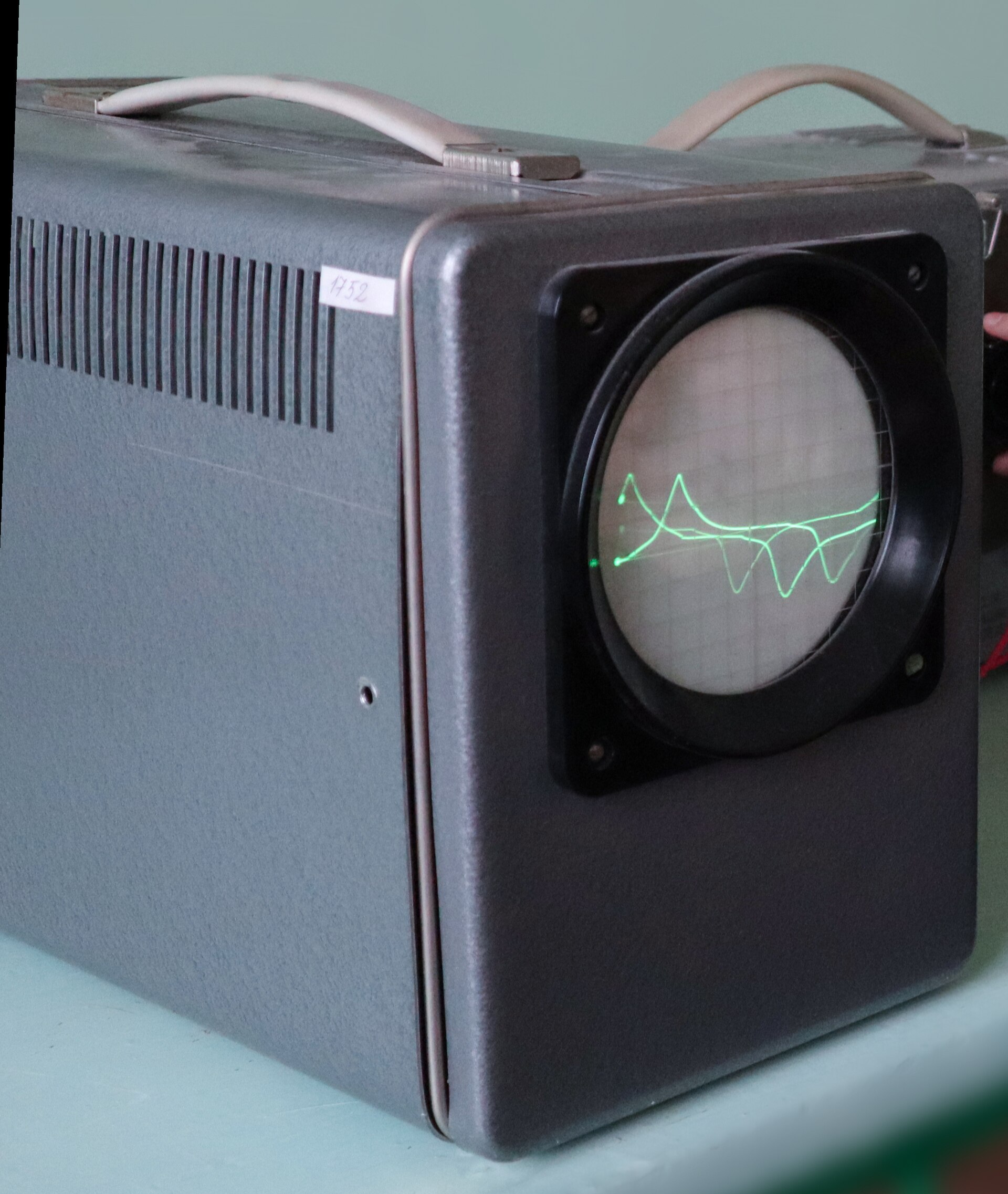 Round CRT oscilloscope screen in a grey steel housing displaying a bright sine wave trace